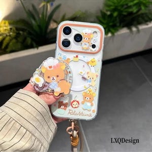 Pode incluir: Uma capa transparente para smartphone com gráficos do Rilakkuma, incluindo um urso com chapéu de chef, um pintinho amarelo e alimentos. A capa tem uma moldura de câmara laranja, um recorte circular e um suporte de telefone a condizer em forma de urso.