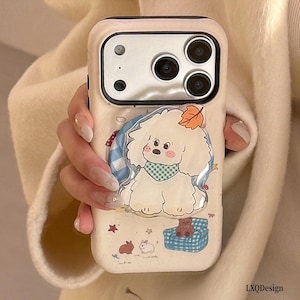 May include: A cream-colored smartphone case featuring a central 3D-style graphic of a fluffy white dog wearing a blue checkered bandana with an orange leaf on its head. The case includes small illustrations of a blue checkered chair, a teddy bear, and rabbits.