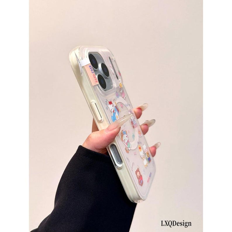 May include: A clear protective phone case featuring a circular magnetic ring and various colorful cartoon character graphics. The case has a light-colored frame, precise cutouts for the camera lenses, and a small rectangular tag labeled KAZD CASE.