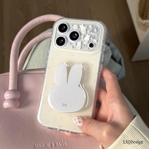 May include: A clear smartphone case featuring a white rabbit-shaped phone grip on the back. The camera area is decorated with small white rabbit figures and star patterns. The case has a subtle star print on the transparent back panel.