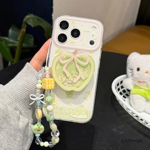 May include: A clear smartphone case featuring a light green apple-shaped pop socket with a pink bow. The case includes a beaded wrist strap with yellow checkered cubes, small apple charms, and clear beads. The design has light green apple graphics.