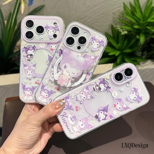 May include: Three clear phone cases featuring purple Kuromi character graphics and accents. Each case has a camera lens cutout with a purple border. One case includes a circular ring stand with a Kuromi figure. The text Kuromi is printed on one case.
