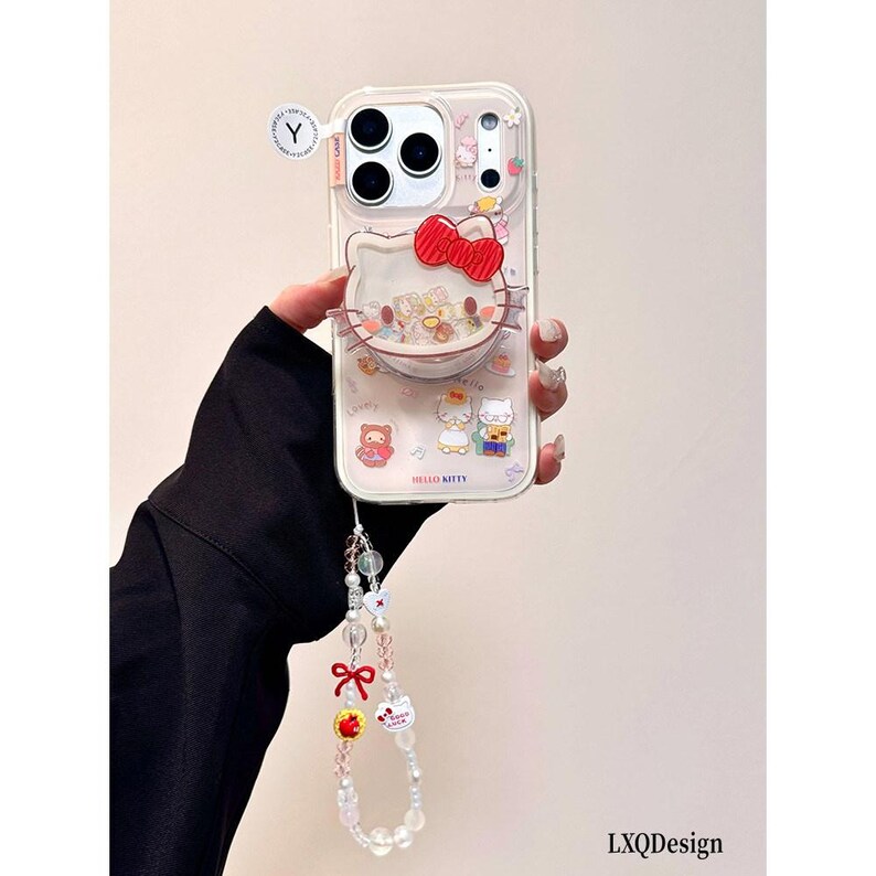 May include: A clear smartphone case featuring a Hello Kitty character design with a red bow, integrated shaker elements, and various small character graphics. The case includes a beaded phone strap with charms, including a red bow and apple.