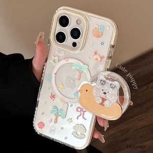 Pode incluir: Uma capa de telemóvel transparente com um anel magnético circular branco e vários ícones pequenos em tons pastel, incluindo palmeiras, conchas, estrelas e um caranguejo. Inclui um suporte circular com um cachorrinho brilhante numa boia.