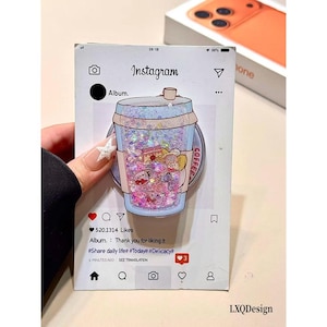 May include: A phone grip featuring a clear, liquid-filled cup design with iridescent glitter and small colorful charms inside. The grip is mounted on a card printed with an Instagram post interface, displaying the text Album, 520,1314 Likes, and hashtags.