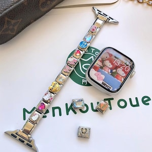 May include: A silver-toned modular watch band featuring various colorful charms, including hearts, stars, and character motifs, paired with a digital smartwatch displaying a cartoon character on its screen. Two additional loose charms are placed nearby.