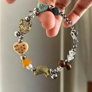 May include: A charm bracelet featuring a variety of beads including a clear cat-shaped bead, a tan heart with star patterns, a yellow mushroom, a crescent moon, and silver-toned metallic spheres. The bracelet is secured with a silver-toned metal clasp.