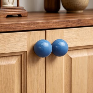 Könnte beinhalten: Zwei glatte, kugelförmige, glänzend blaue Schrankknöpfe sind an der Vorderseite von hellen Holzschranktüren befestigt. Die Knöpfe haben eine solide, abgerundete Form und eine reflektierende Oberfläche, die mit der natürlichen Holzmaserung kontrastiert.