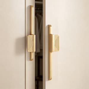 May include: Two vertical gold-toned metal cabinet handles mounted on light-colored doors. Each handle features a long, smooth bar section and a central rectangular grip area decorated with a series of concentric, stepped square ridges.