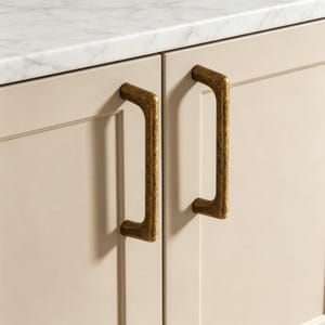 Può includere: Due maniglie verticali per armadietti in ottone con una finitura testurizzata e martellata. Le maniglie sono montate su ante di armadietti color beige chiaro con design a pannello, posizionate sotto un piano in marmo bianco.