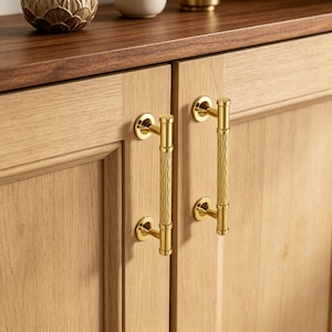 May include: Two vertical brass cabinet handles with a textured, ribbed centre grip and smooth circular mounting plates. The handles are attached to light-toned wooden cabinet doors, featuring a polished metallic finish and a cylindrical design.
