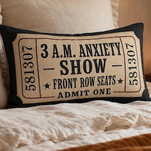 May include: A rectangular black and cream throw pillow featuring a graphic design of a vintage ticket. The text reads 3 A.M. ANXIETY SHOW, FRONT ROW SEATS, and ADMIT ONE, with the number 581307 printed vertically on both sides of the ticket design.