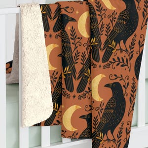 May include: A plush throw blanket featuring a repeating pattern of black crows, golden crescent moons, and dark leafy branches on a burnt orange background. The reverse side of the blanket is made of a thick, cream-coloured sherpa fleece material.