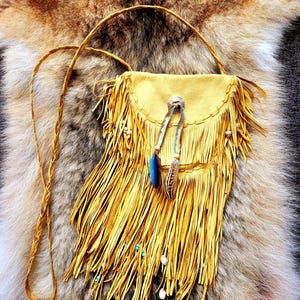 Peut inclure: Un sac bandoulière en cuir jaune moutarde avec de longues franges, une bandoulière tressée et un rabat. Le sac est orné d'un concho en métal argenté, de deux cordons perlés avec des plumes et de petites perles blanches et bleues.