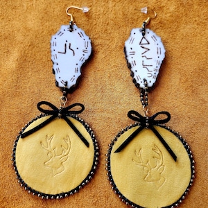 May include: A pair of dangle earrings featuring a white, irregular-shaped top piece with black etched symbols, connected by black beads to a circular yellow leather pendant embossed with a deer head and accented with a small black ribbon bow.