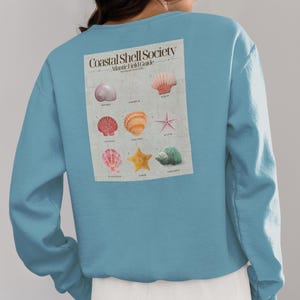 May include: A light blue long-sleeve sweatshirt featuring a rectangular graphic on the back titled Coastal Shell Society Atlantic Field Guide. The print displays nine illustrations of various seashells and starfish in shades of pink, orange, and green.