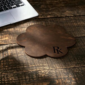 May include: A dark brown, flower-shaped leather coaster featuring the letters RK embossed in black on the bottom right side. The coaster has a smooth, matte finish and rounded edges, providing a distinct shape for resting beverages or small items.
