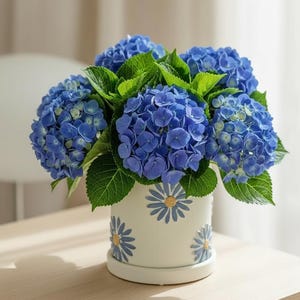 May include: A bouquet of vibrant blue hydrangea flowers with lush green leaves arranged in a white ceramic planter. The planter features a repeating pattern of blue daisy-like flowers with yellow centres embossed around its cylindrical exterior.