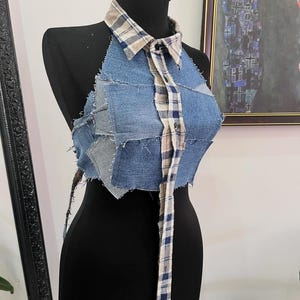 Może przedstawiać: Top typu halter wykonany z patchworkowego niebieskiego dżinsu z postrzępionymi krawędziami. Ubiór posiada kołnierzyk w beżowo-granatowo-białą kratę oraz długi, wąski pasek materiału w kratę zwisający w dół przez środek przedniej części.