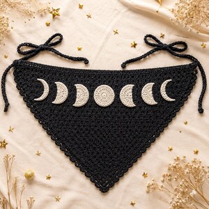 May include: A black triangular crochet bandana featuring a series of seven cream-colored moon phases appliqued across the top. The accessory has a scalloped edge and long crocheted ties at the two upper corners for securing the garment in place.