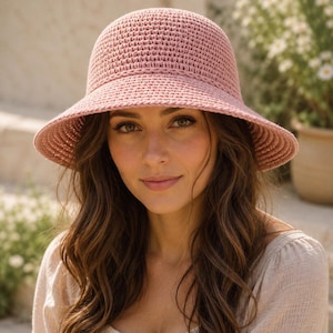 Può includere: Un cappello a secchiello all'uncinetto color rosa polvere, caratterizzato da una calotta arrotondata e una tesa inclinata verso il basso. Il cappello è realizzato con un punto denso e uniforme, creando una superficie testurizzata e omogenea.