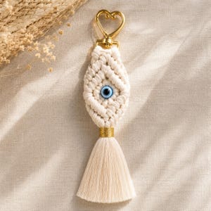 May include: A macrame keychain featuring an off-white knotted cord design with a central blue and white evil eye bead. The piece is attached to a gold-toned heart-shaped metal clasp and finished with a long, straight off-white fiber tassel.