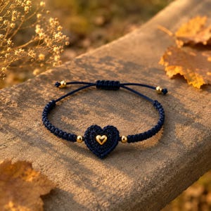May include: A navy blue cord bracelet featuring a central crocheted heart charm with a small gold-toned heart accent in the middle. The bracelet includes four small gold-toned spherical beads and an adjustable sliding knot closure mechanism.