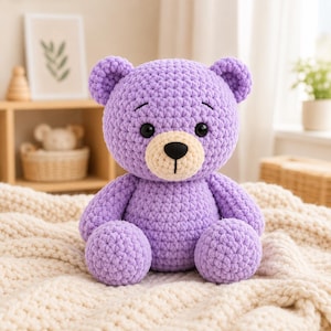 May include: A handmade crochet teddy bear crafted from soft, lavender-coloured yarn. The plush toy features a round head with small ears, a cream-coloured muzzle, a black stitched nose, and two dark, round plastic eyes. It sits in an upright position.