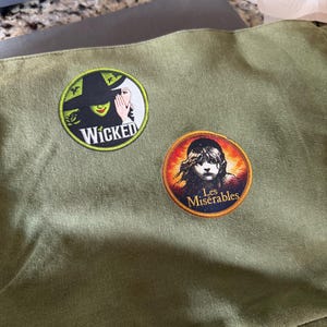 May include: An olive green canvas bag featuring two circular embroidered patches. The top patch displays a green-skinned figure in a black hat with the text Wicked. The bottom patch shows a young person with the text Les Misérables.