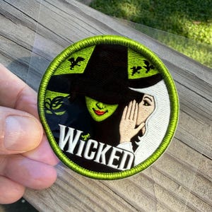 May include: A circular embroidered patch featuring a green-skinned figure in a black pointed hat and a white-skinned figure whispering, with the word Wicked in white text at the bottom and small black flying monkey silhouettes on a lime green background.