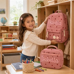 May include: A pink corduroy backpack and a matching lunch bag, both featuring a strawberry pattern and the name Isabella embroidered in white script. The backpack has two small flower appliqués on the front pocket and white zipper pulls.