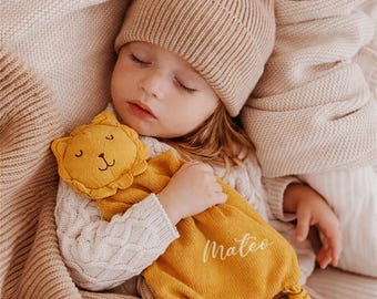 Könnte beinhalten: Eine senfgelbe Strick-Schnuffeltuch mit einem Löwenkopf mit gestickten Gesichtszügen und dem in weißer Schreibschrift aufgestickten Namen Mateo auf der Vorderseite, mit verknoteten Ecken und einer weichen, strukturierten Stoffbeschaffenheit.