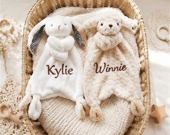 Könnte beinhalten: Zwei Plüsch-Schmusetücher mit Tierköpfen. Der weiße Hase hat schwarz-weiß gestreifte Innenohren und den in Braun gestickten Namen Kylie. Das beige Lamm hat einen strukturierten Fleece-Körper und den in Braun gestickten Namen Winnie.
