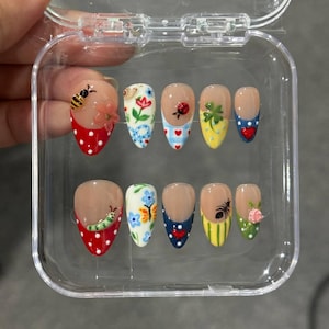 May include: A set of ten press-on nails in a clear plastic case, featuring almond-shaped tips with various designs including a bee, ladybug, caterpillar, butterfly, flowers, hearts, polka dots, and stripes in vibrant red, blue, yellow, and green.