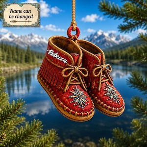 May include: A pair of red hanging moccasin ornaments with tan laces, fringe detailing, and a beaded star pattern on the toe. The side of the left moccasin features the name Rebecca in white script. A sign above reads Name can be changed.
