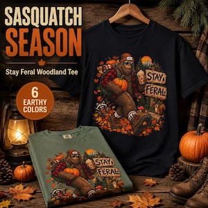May include: A black and a sage green t-shirt featuring a graphic of a Sasquatch wearing a beanie, sunglasses, and a red plaid shirt, holding a pumpkin and a drink. The design includes the text Stay Feral, autumn leaves, a raccoon, and a squirrel.