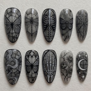 May include: A set of ten almond-shaped press-on nails featuring a grey and white marble base with black line art designs including a butterfly, dragonfly, ribcage, moth, spiderweb, smoke, gothic arch, skeleton hand, flowers, and a crescent moon.