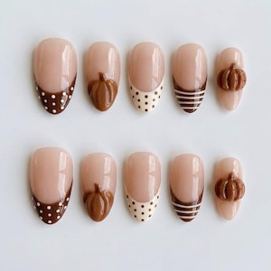 May include: A set of ten press-on nails featuring a nude base with brown accents. Designs include brown tips with white polka dots, solid brown raised pumpkin shapes, nude tips with brown polka dots, brown and white horizontal stripes, and solid nude.