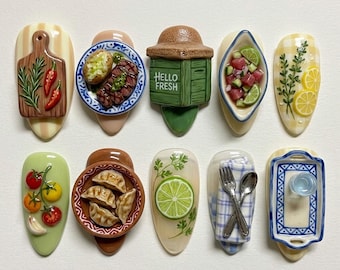 May include: A set of ten decorative ceramic magnets featuring miniature food items including a cutting board with herbs, a steak dinner, a Hello Fresh crate, a bowl of ceviche, lemon slices, tomatoes, dumplings, a lime slice, cutlery, and a tray.