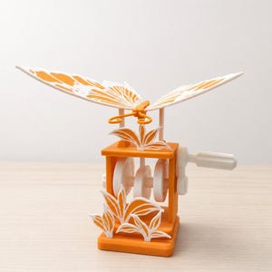 May include: A mechanical orange and white butterfly automaton featuring a hand crank on the side. The butterfly has orange wings with white patterns, mounted on a central orange frame with white internal gears and decorative white leaf cutouts.