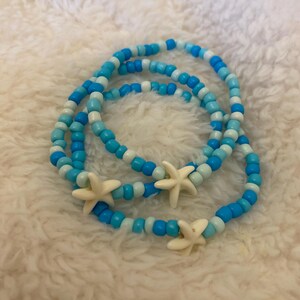 May include: Three circular beaded bracelets featuring a repeating pattern of light blue, medium blue, and white spherical beads. Each bracelet is accented with a single cream-colored, five-pointed starfish charm attached to the strand of beads.