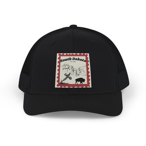 May include: A black trucker hat featuring a rectangular graphic on the front panel. The graphic resembles a postage stamp with a red border, displaying the text South Dakota EST 1889, Mount Rushmore, a flying pheasant, and a black bison silhouette.