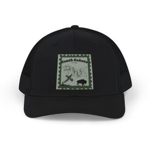 Puede incluir: Una gorra de camionero negra con un gráfico cuadrado en el panel frontal. El gráfico se asemeja a un sello postal con borde verde, que representa el Monte Rushmore, un ave en vuelo y un bisonte, con el texto South Dakota y EST. 1889.