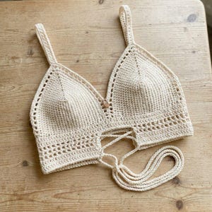 May include: A cream-colored crochet crop top featuring a triangular bralette design with two shoulder straps, a central wooden bead detail, and a decorative open-stitch pattern along the bottom edge with long, adjustable tie-up strings.