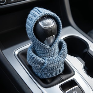 May include: A light blue crocheted hoodie cover designed to fit over a car gear shift knob. The miniature garment features a hood, long sleeves, and a front pouch pocket, crafted with a textured knit pattern that fits snugly around the black gear shifter.
