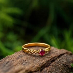 Può includere: Un anello in oro con una pietra preziosa rubino rosso scuro di forma rotonda al centro. La fascia dell'anello presenta un dettagliato motivo a foglie in rilievo che si estende dall'incastonatura centrale lungo entrambi i lati del cerchio.