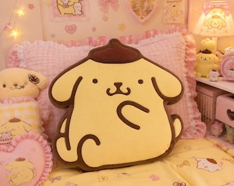 Può includere: Un cuscino decorativo in peluche a forma del personaggio Pompompurin, caratterizzato da un corpo giallo chiaro con un basco marrone scuro, tratti del viso marrone scuro e un braccio curvo. Il cuscino ha una consistenza morbida su un cuscino rosa.