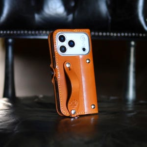 May include: A tan leather phone case with a built-in hand strap and silver-toned metal rivets. The case features a cutout for a triple-lens camera system and a small embossed logo with a clover symbol and the text Ricky's on the strap.