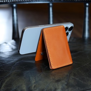 May include: A tan leather magnetic phone wallet stand attached to the back of a silver smartphone. The wallet features visible yellow stitching along the edges and a small metal snap button on the side, functioning as a kickstand for the device.