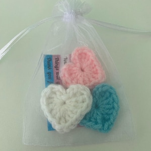 May include: A sheer white organza drawstring bag containing three small crocheted hearts in light pink, white, and light blue. A small rectangular card is visible inside the bag with the printed text: You. Things will g. Thank you fo.
